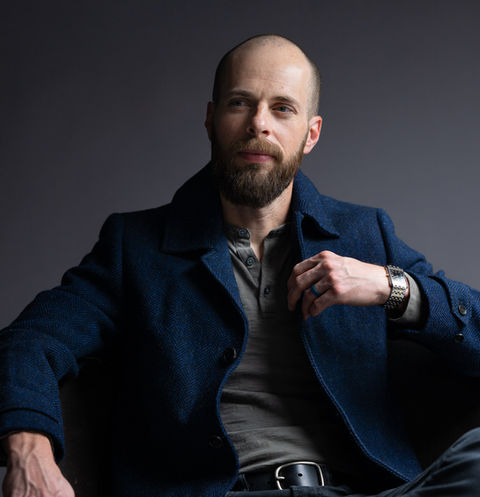 Dramatic studio portrait of a bearded man in a textured blue coat seated in a chair, styled for creative headshots and editorial profiles.