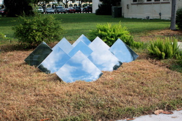A sculpture of squares in a garden area, they appear to be reflecting the sky, which looks fascinatingly odd seeing as they are grounded in mulch.