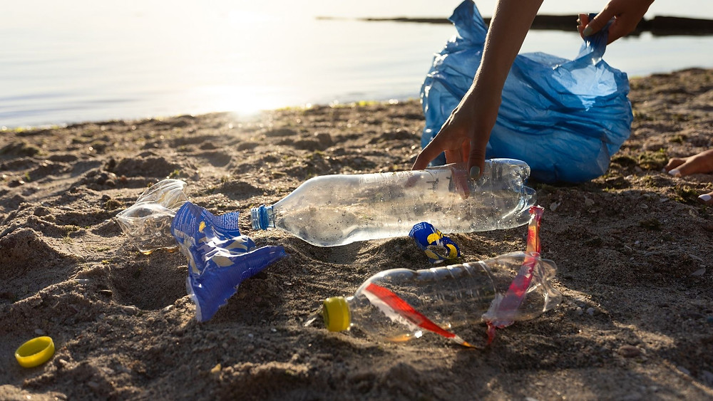 A hand reaches down to pick up plastic bottles on a beach. The other visible hand carries a trashbag.