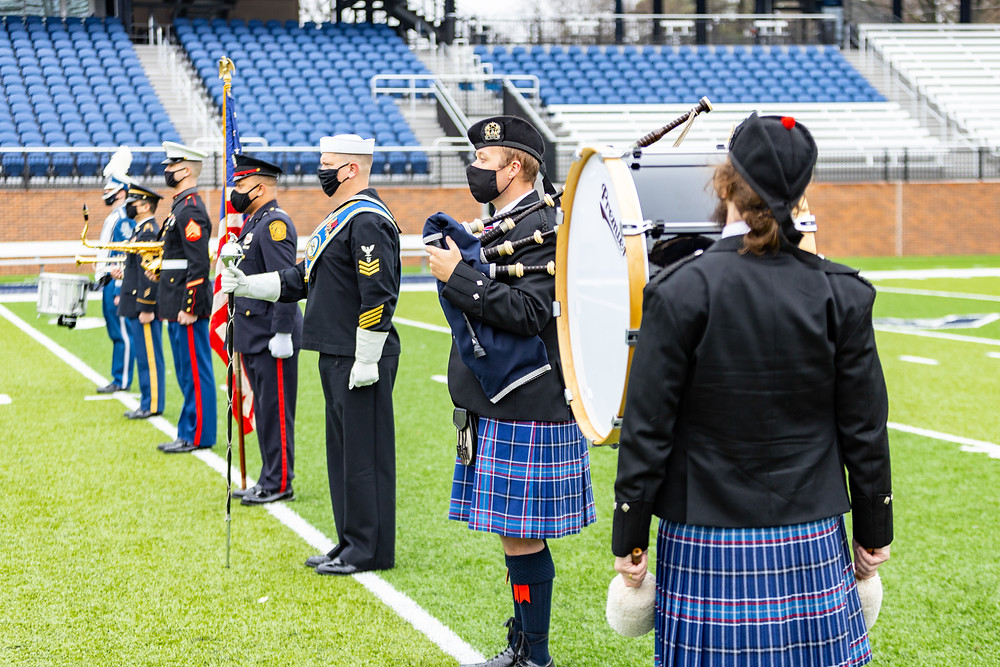 A line of drummers, pipers, brass musicians, a flag bearer and a drum major standing at attention on a football field for the Tattoo press conference. They are wearing their military uniforms, or the Tattoo tartan kilts (depending on what would be appropriate)