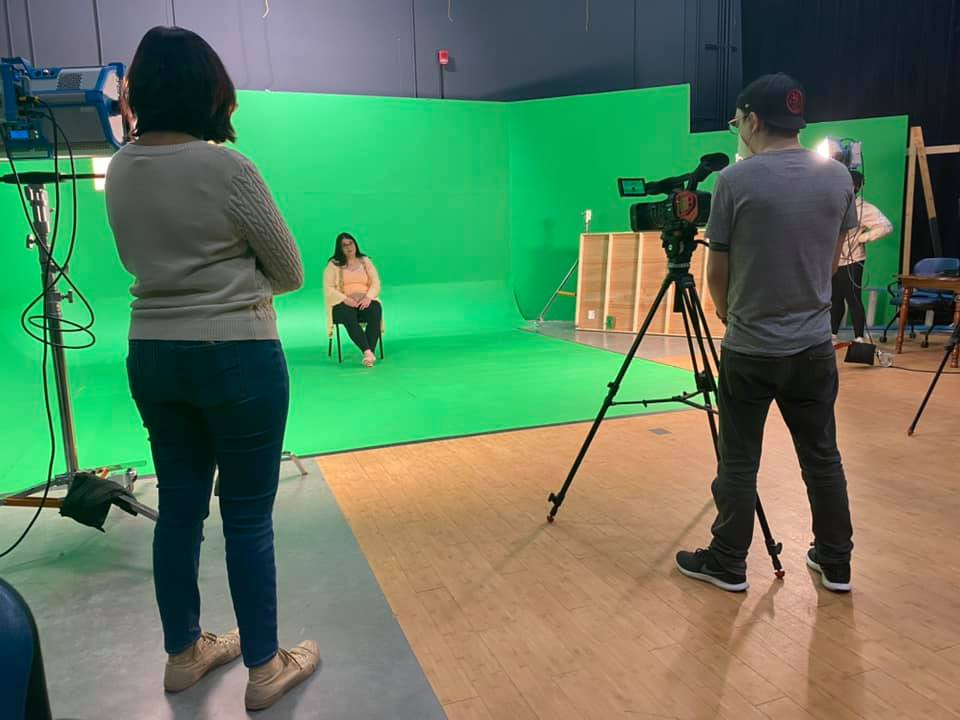 Aliki sits on a chair in front of a green screen, with a film crew surrounding her, all socially distanced and wearing masks