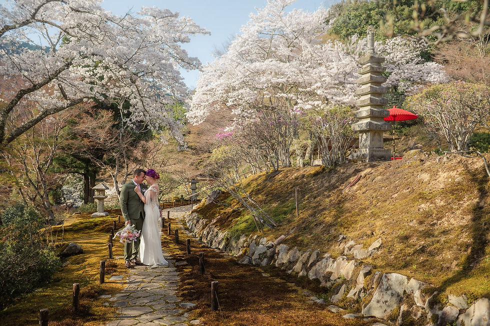 A couple embraces on a stone path in a Japanese garden with cherry blossoms and a red umbrella. The mood is romantic and serene.