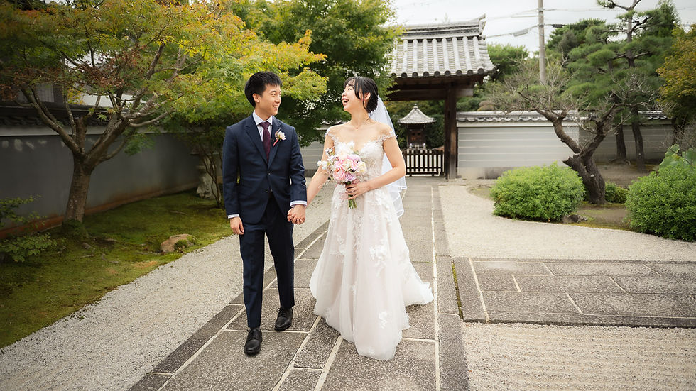 Bride and groom smiling, holding hands in a Japanese garden. She wears a white gown, he a dark suit. Lush greenery and traditional gate.