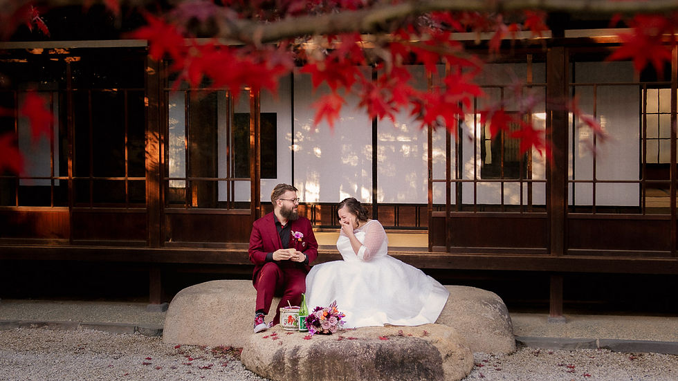 A couple sits on stones under red leaves, the man in a burgundy suit, the woman in a white dress, laughing in a serene Japanese setting.