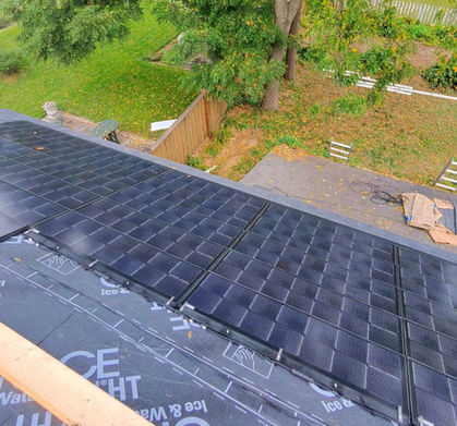 Close-up view of dark, integrated solar shingles being installed on a roof over an 'Ice & Water Shield' underlayment, with a wooden fence and grassy yard visible in the background.