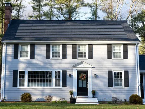 Dark solar shingles on a sloped roof have melted the surrounding snow, creating clear runoff paths and revealing the shingles beneath. The rest of the roof is still covered in white snow, highlighting the panels' heat absorption. A snowy rooftop scene under blue sky with pine trees in the background.