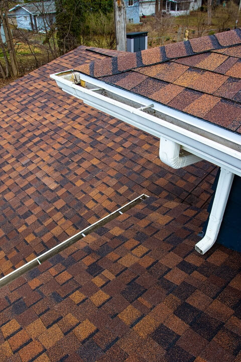Close-up, high-angle view of a roof showing multi-toned brown asphalt shingles, a clean white gutter, and downspout.