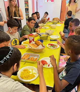 teens sitting around a long table with food for them to serve themselves, hot dogs, corn, tomatoes, cucumbers and other vegetables