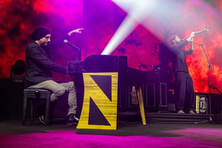 Idan Raichel plays piano on stage while another performs with a trumpet, illuminated by colorful lights.