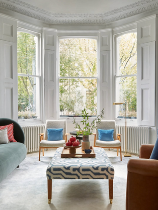 Bright and elegant living room with a large bay window and detailed decorative cornicing. The space features a soft blue velvet sofa, two mid-century style armchairs with blue cushions, a patterned ottoman used as a coffee table with a wooden tray and decorative items, and a warm tan armchair. Natural light floods the room, highlighting the classic white panelled walls and tall sash windows.