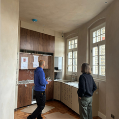 On-site image of a renovation in progress, with two people discussing kitchen installation details. Custom cabinetry, arched sash windows, and protective flooring are visible.
