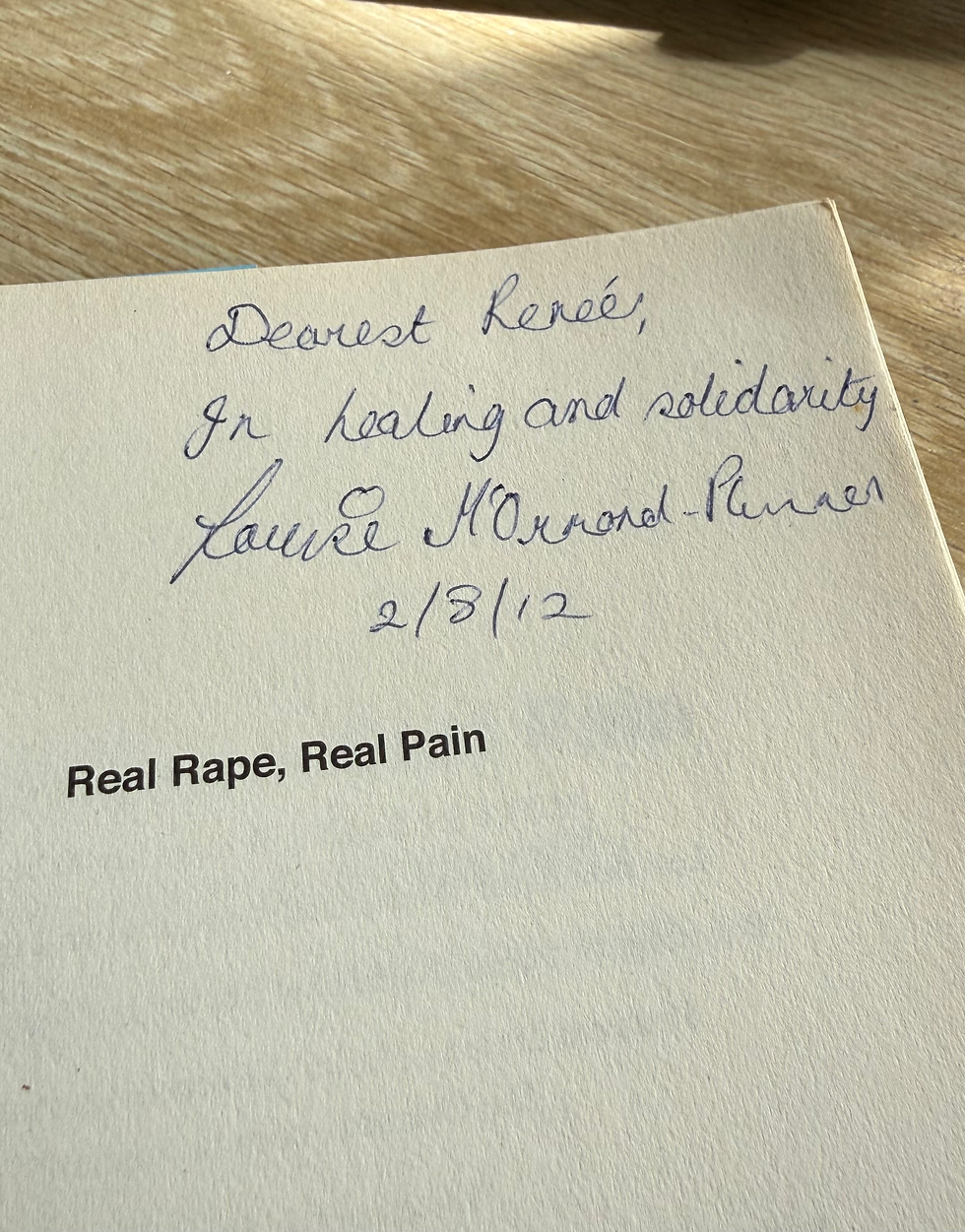 Book page on a wooden table with handwritten note: "Dearest Renée, In healing and solidarity" above text "Real Rape, Real Pain." Warm lighting.
