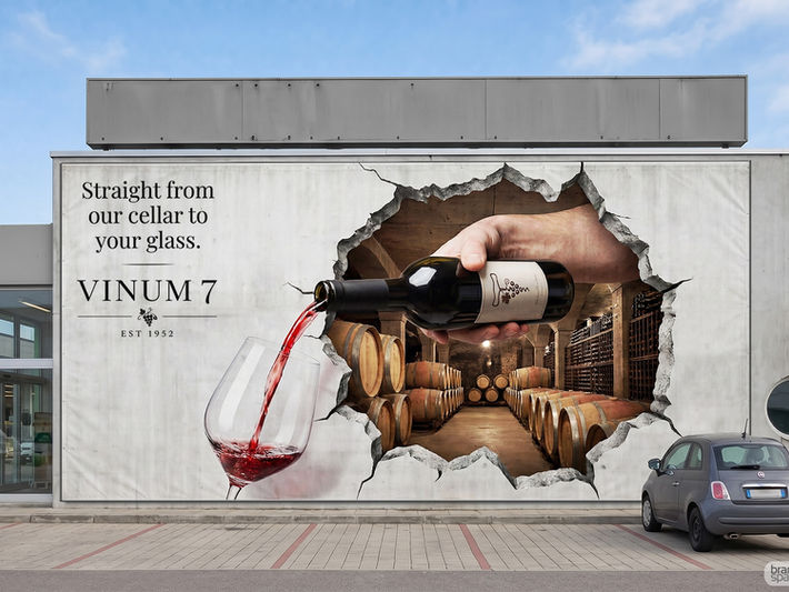 A massive 3D building wrap designed by Brandspace3D on a concrete commercial building, creating a realistic optical illusion of a giant hole breaking open to reveal a wine barrel cellar. A giant hand pours red wine from a 'VINUM 7' bottle into a large glass, alongside the text "Straight from our cellar to your glass." A small grey car is parked next to it.