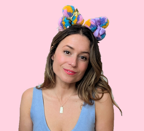 A woman with brunette hair and a blue top wears a pom-pom head piece on her head, tilted slightly. She has red lipstick on.