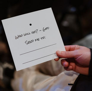 A person holding a square piece of paper that states Who Will Go? God send me to: