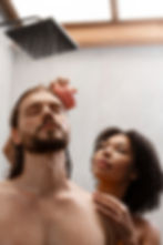Man with closed eyes in shower, his hair being scrubbed with a pink brush by a woman. Water streams down, creating a calming mood.