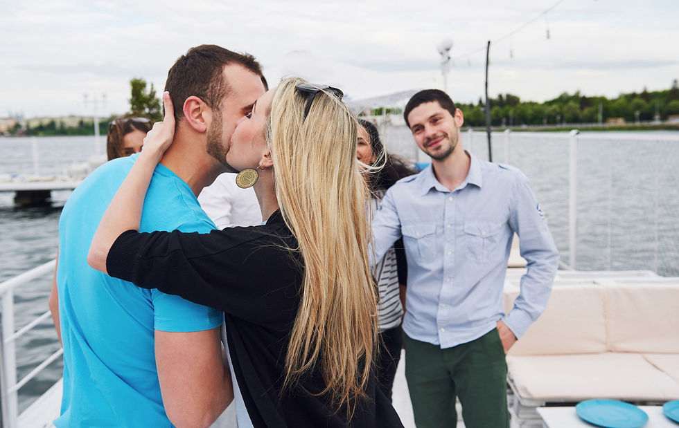 A couple kisses on a boat, surrounded by friends. The setting is a calm river, and everyone appears happy. The man wears a blue shirt.