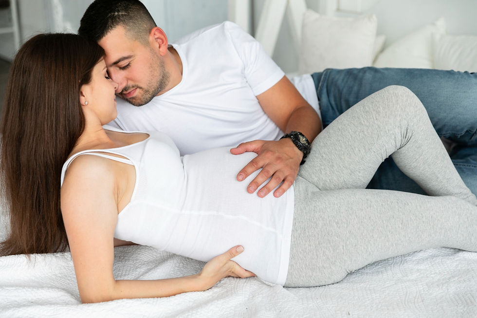 Man and woman lying on bed, touching woman's pregnant belly. Both wear white tops, man in jeans. Cozy, intimate setting with pillows.