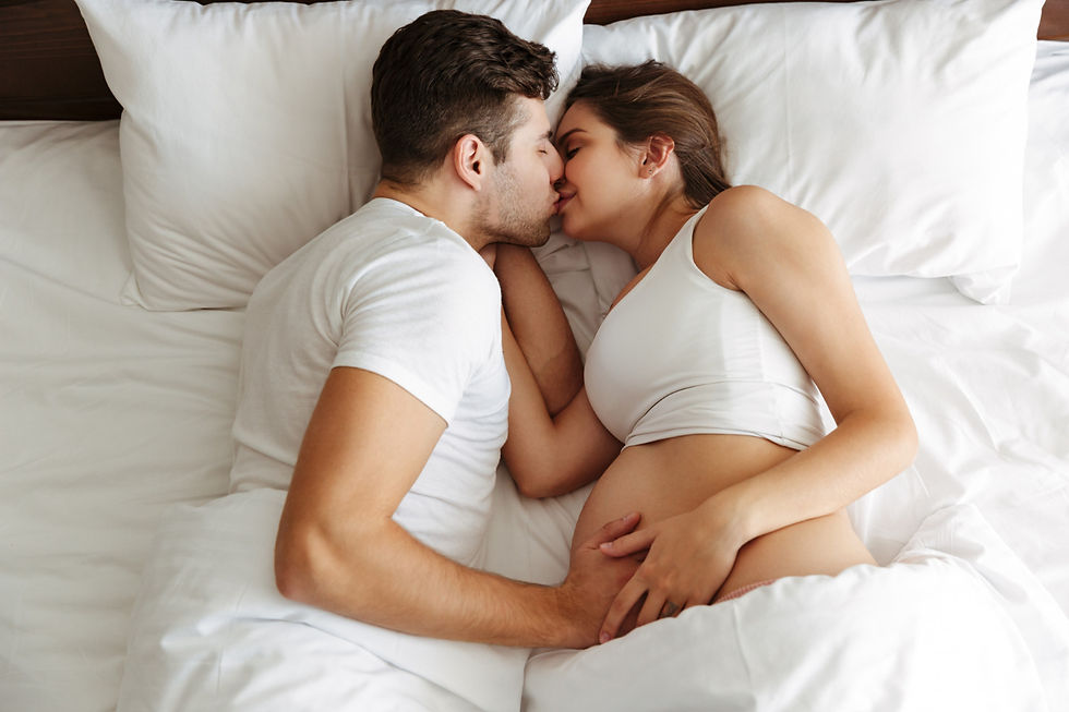 A couple in white clothing lies in bed, embracing and smiling. The woman is pregnant. The setting is cozy, with white pillows and bedding.
