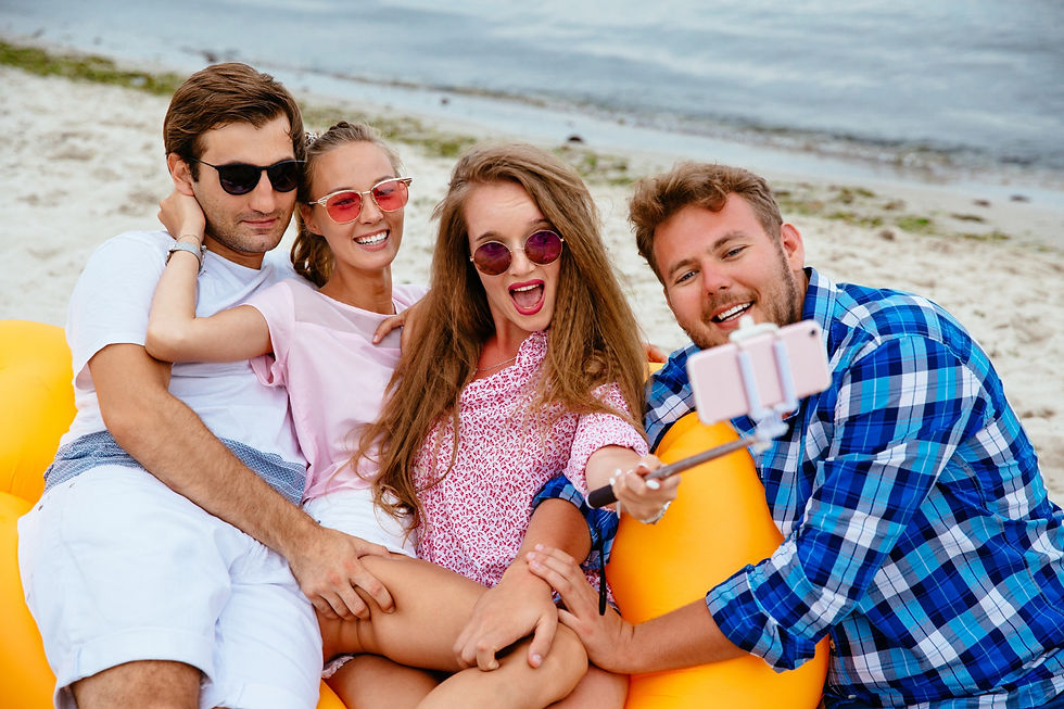 Four friends taking a beach selfie on a yellow inflatable, wearing sunglasses, smiling and happy. Ocean and sand in the background.