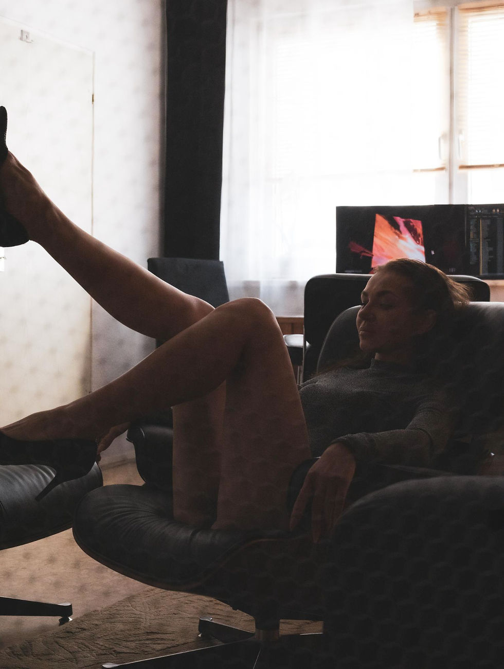 A woman relaxes on a black chair in a dimly lit room, with one leg raised. A computer displaying abstract art is in the background.