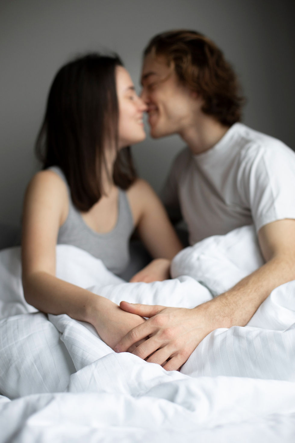 A couple sits closely under white sheets, smiling and leaning in for a kiss in a cozy, gray-walled room. Their hands are gently intertwined.