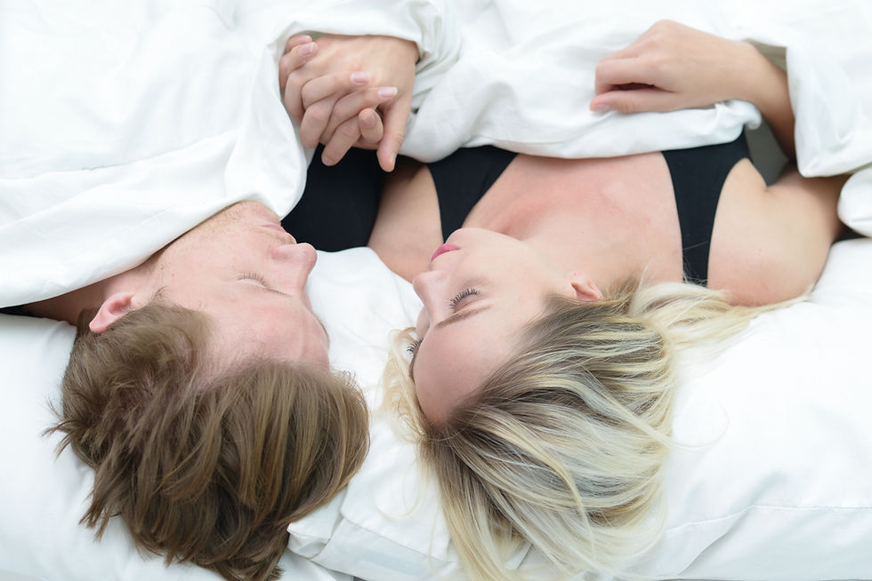 Couple lying in bed under white sheets, holding hands. They're facing each other with calm expressions, on white pillows in a serene setting.