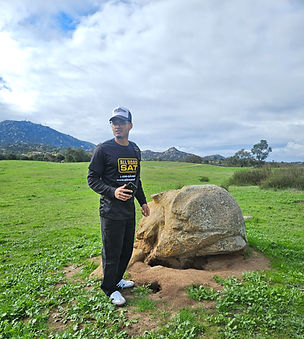 Man in black clothes stands by large rock, green field, cloudy sky.