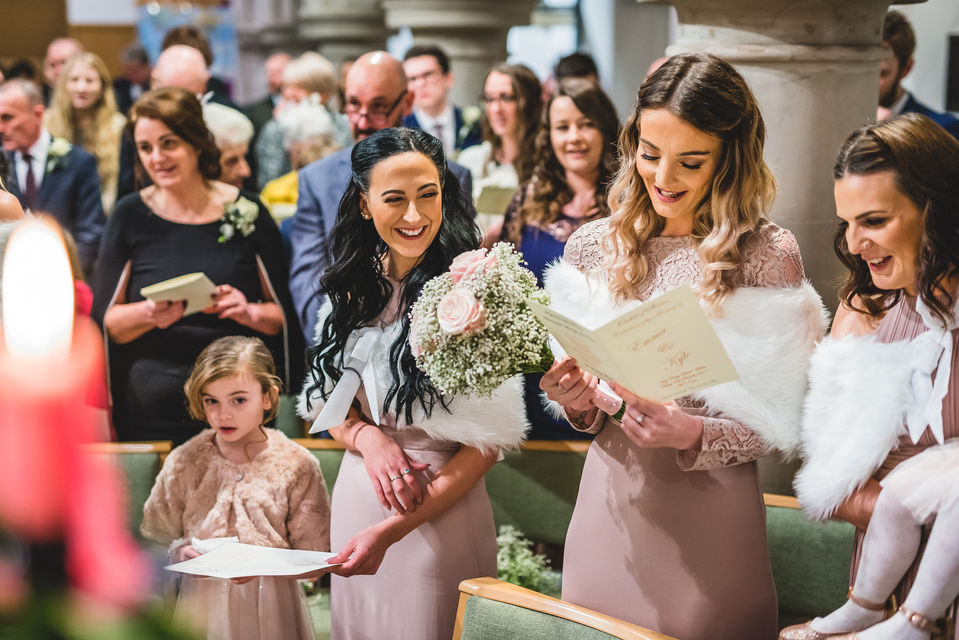 Brides maids wearing long pink dressed and holding pink peonies laugh as they sing in church at a wedding, image captured by wedding photographer Aaron Scott Richards in Birmingham