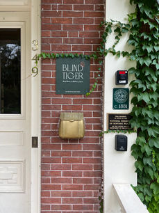 Vertical close-up of brick wall with green “Blind Tiger” guest house sign, brass mailbox, and ivy detail.
