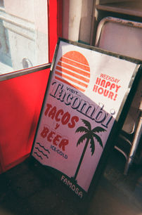Slanted Tacombi signboard reading “Weekday Happy Hour! Tacos y Beer,” placed beside a red interior wall.