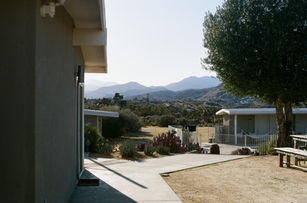 Outdoor scene with white buildings, desert plants, and mountains in the background, viewed from a shaded patio.