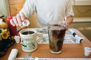 A person pouring creamer into a mug of coffee at a diner table, with a tall iced coffee next to it and retro condiments in the background.