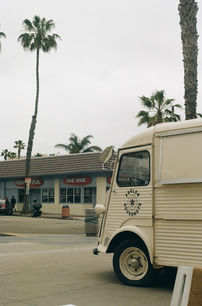 Vintage white Parlor Doughnuts truck parked in front of a strip mall with palm trees and a cloudy sky.