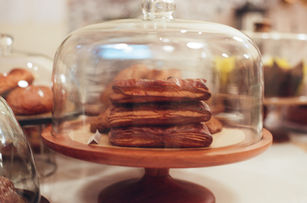 Close-up of rectangular pastries stacked under a glass cloche on a wooden pedestal stand, surrounded by other baked goods.