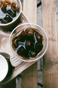 Overhead close-up of two iced coffees with ice cubes visible, sitting in a cardboard drink carrier on a wooden table.