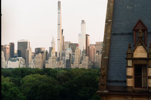 City skyline view with tall modern skyscrapers rising above lush park trees, framed by classic stone building rooftop in foreground.