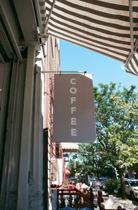Outdoor coffee shop sign reading "COFFEE" in white letters on a neutral background, under a striped awning with blue sky and trees behind it.