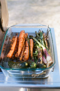 Glass dish filled with grilled carrots, scallions, and other charred vegetables, sitting on a metal outdoor table.