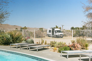 Poolside scene with modern lounge chairs and desert plants, looking out toward a parked white van and Joshua trees in the background.