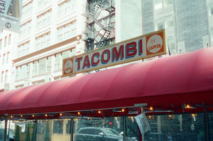 Wide shot of city street corner with bicycle lane, large TACOMBI mural painted on building, and warm afternoon light.