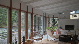 Bright open-concept living room with white ceiling beams, floor-to-ceiling windows, plants, and mid-century style chairs.