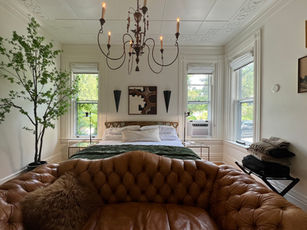 Spacious guest room featuring white walls, tall windows, vintage chandelier, and brown leather sofa at the foot of a bed.