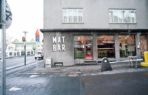 Street-facing view of minimalist gray building with bold white “MAT BAR” text, large windows showing cafe and bar interior.