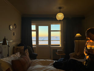 Dimly lit guest room with bed and lounge chairs, snow-covered view through window, person standing near lamp.