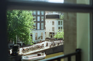 Window-framed view of canal, bicycles, and classic Dutch buildings with leafy trees.