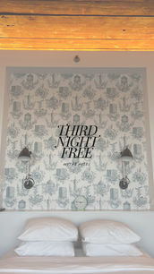 Darling and minimal guest room with patterned wallpaper, two sconces, and white pillows stacked on twin beds, featuring ‘Third Night Free’ promotional text.