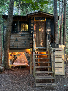 Two-level wooden treehouse structure with front steps, fairy lights, and hammock bed below, surrounded by pine trees.