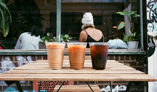 Iced coffee trio on wood table outside Ludlow Coffee Supply, café window and seated person with white beanie in background.