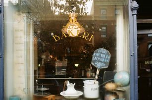 Bakery window with gold “BAKERI” lettering, cake stand, flour jars, and chalkboard hours sign reflected alongside warm interior lights.